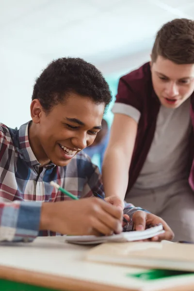 Sonriente colegial afroamericano escribiendo en libro de texto y compañero de clase parado detrás - foto de stock