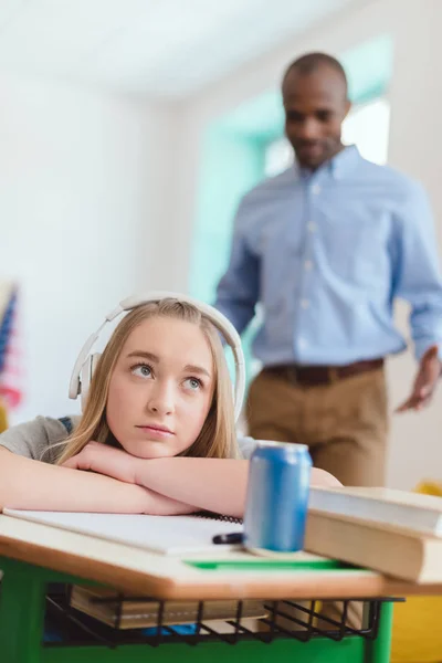Lycée adolescent femme écouter de la musique dans les écouteurs pendant la leçon et professeur afro-américain debout derrière — Photo de stock