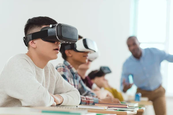 Visão lateral de escolares multiculturais usando fones de ouvido de realidade virtual e professor falando em pé atrás — Fotografia de Stock