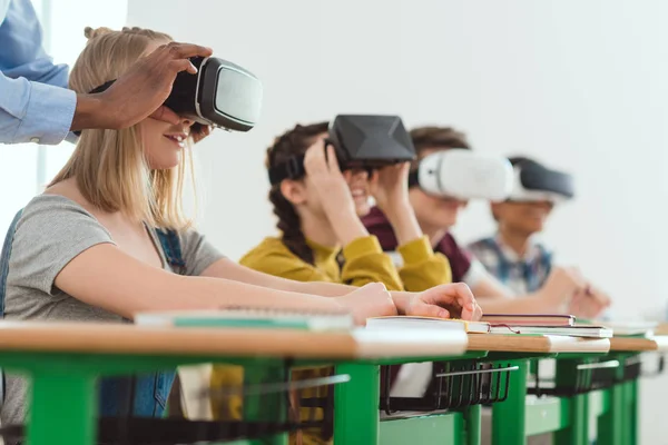 Imagen recortada del profesor afroamericano ajustando los auriculares de realidad virtual a la colegiala adolescente con compañeros sentados detrás - foto de stock