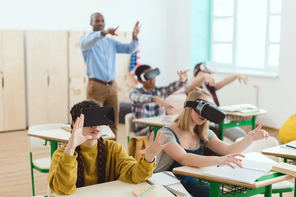Estudantes adolescentes do ensino médio usando fones de ouvido de realidade virtual e professor de gestos em pé atrás em sala de aula — Fotografia de Stock