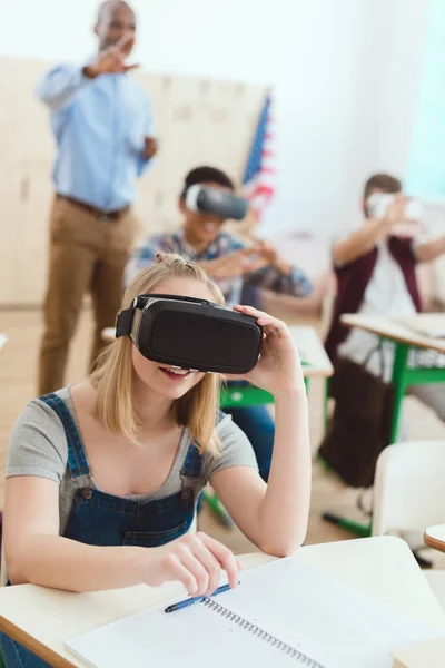 Teenage schoolgirl using virtual reality headset with male classmates and teacher behind — Stock Photo