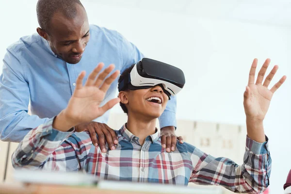 Sonriente estudiante adolescente de secundaria usando auriculares de realidad virtual y profesor sosteniendo sus hombros - foto de stock