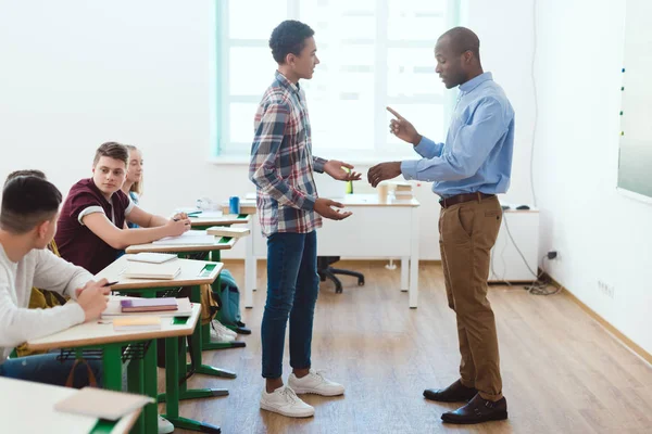 Side view of african american teacher pointing at wristwatch to latecomer schoolboy in classroom — Stock Photo