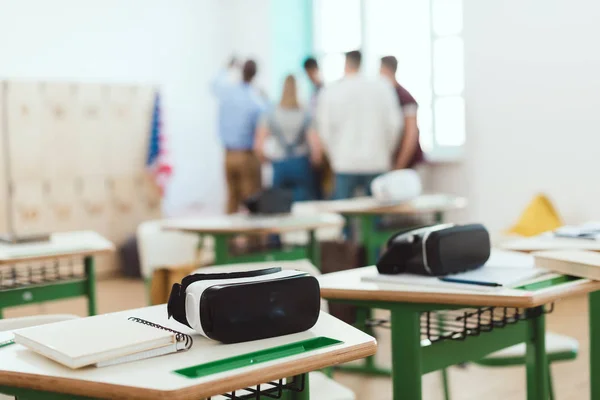 Fones de ouvido de realidade virtual em mesas com professores e alunos do ensino médio em pé atrás em sala de aula — Fotografia de Stock