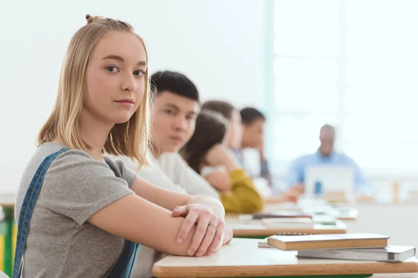 Portrait of caucasian schoolgirl sitting at desk with classmates and teacher behind — Stock Photo