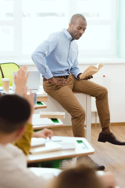 Profesora afroamericana sentada en la mesa y leyendo libro a escolares con los brazos en alto en el aula - foto de stock