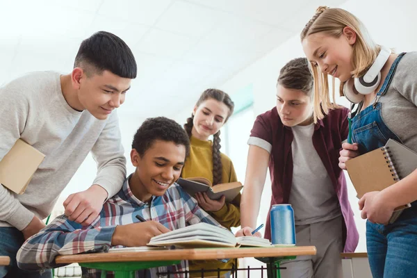 Sonriente afroamericano colegial escribir en cuaderno mientras sus compañeros de clase de pie alrededor - foto de stock