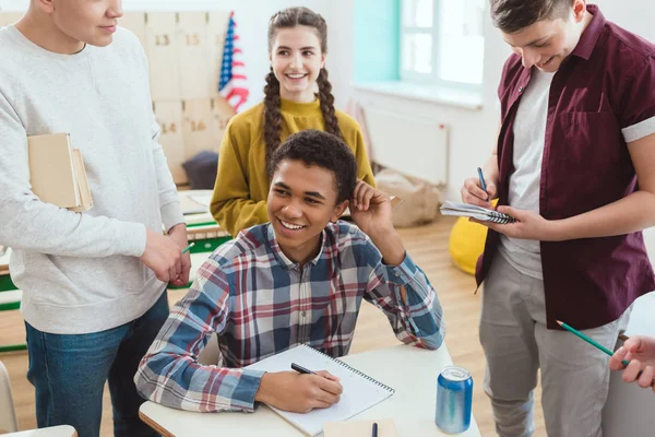 Happy african american schoolboy writing in notebook while his classmates standing around — Stock Photo