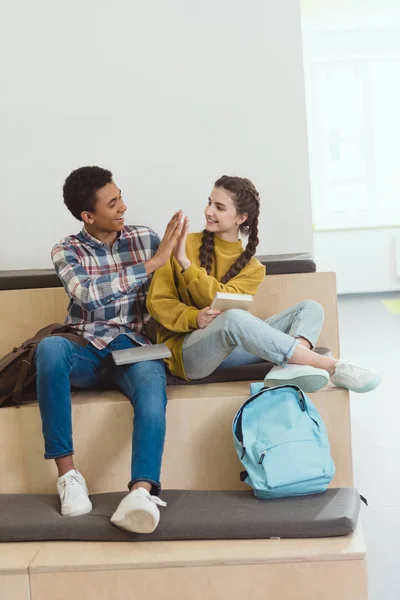 Estudiantes de secundaria pareja sentados juntos en el pasillo de la escuela y dando cinco - foto de stock