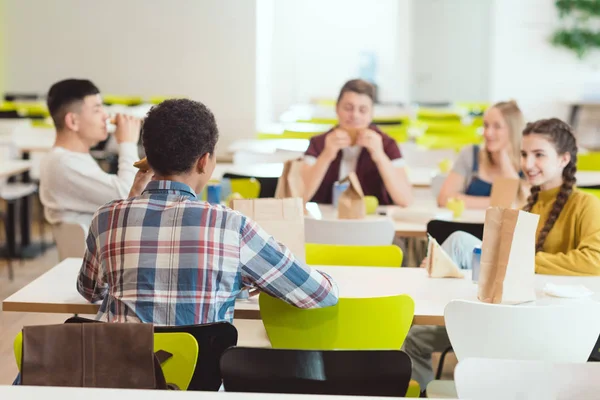 Groupe d'élèves adolescents bavardant tout en prenant le déjeuner à la cafétéria de l'école — Photo de stock