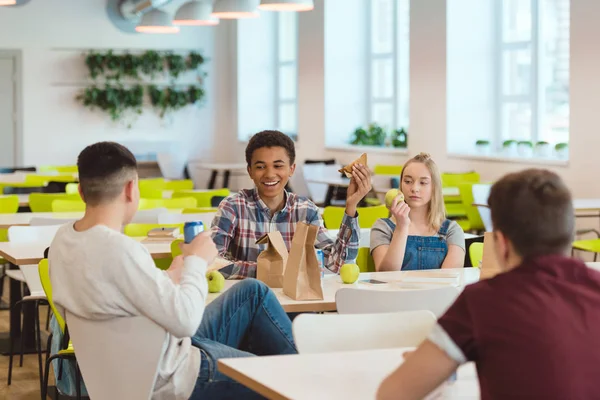 Feliz grupo multiétnico de estudiantes de secundaria charlando mientras almuerzan en la cafetería de la escuela - foto de stock