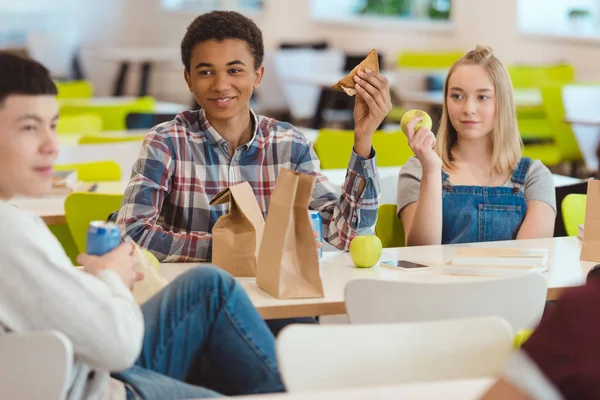 Grupo multiétnico de estudiantes de secundaria charlando mientras almuerzan en la cafetería de la escuela - foto de stock