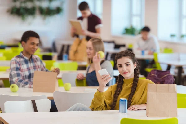 Souriant adolescent écolière assis à l'école cafétéria avec des camarades de classe et regardant caméra — Photo de stock