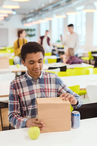 Colegial afroamericano abriendo bolsa de almuerzo en cafetería escolar - foto de stock