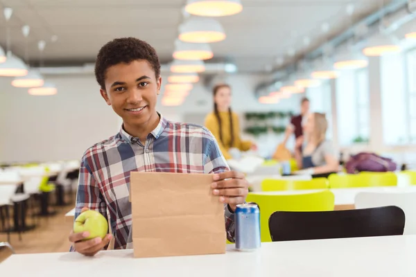 Colegial afroamericano con manzana y bolsa de almuerzo en cafetería de la escuela - foto de stock