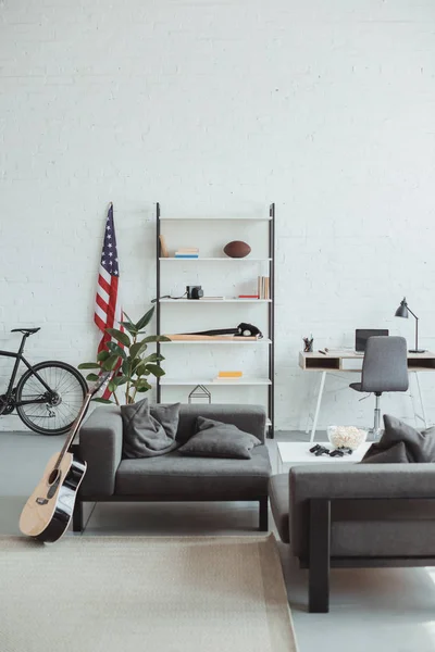 Interior de la moderna sala de estar con bicicleta, guitarra, bandera americana, ordenador portátil, estantes y sillones - foto de stock