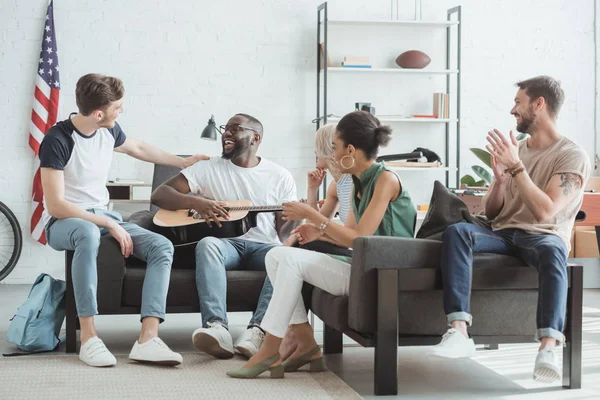 Group of multiethnic smiling young people playing guitar in living room — Stock Photo