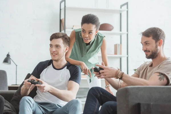 Joven afroamericana mujer viendo dos hombres jugando videojuego con joysticks en las manos - foto de stock