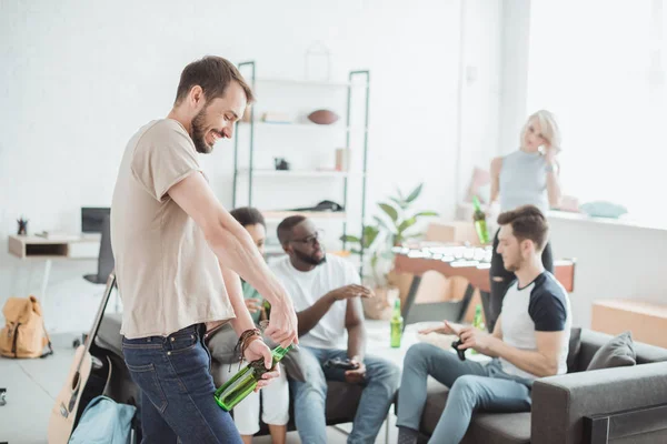 Seitenansicht eines jungen Mannes, der mit einem Korkenzieher eine Bierflasche öffnet und sich dahinter mit Freunden unterhält — Stockfoto