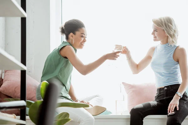 Junge lächelnde multiethnische Freundinnen, die Gläser auf dem Fenstersims klirren lassen — Stockfoto