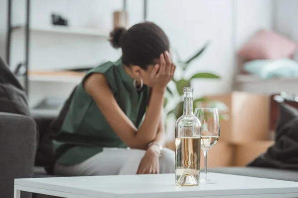 Primo piano colpo di vetro con bottiglia di vino sul tavolo e frustrata giovane donna seduta in poltrona — Foto stock