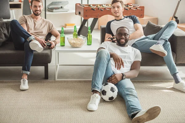 Jovem negro homem sentado no tapete com bola entre dois amigos jogar vídeo game com joysticks em mãos — Fotografia de Stock