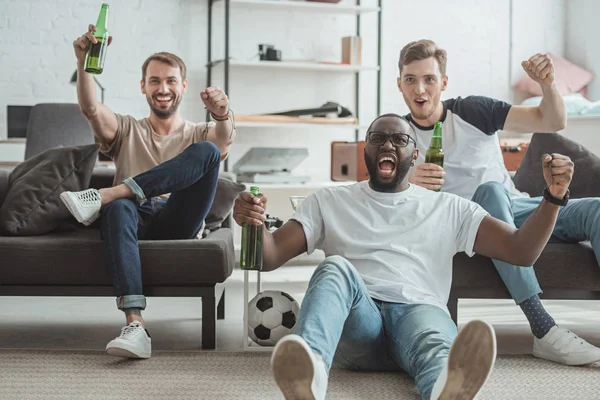 Jóvenes felices amigos masculinos multiculturales viendo el partido de fútbol y celebrando con botellas de cerveza en las manos - foto de stock