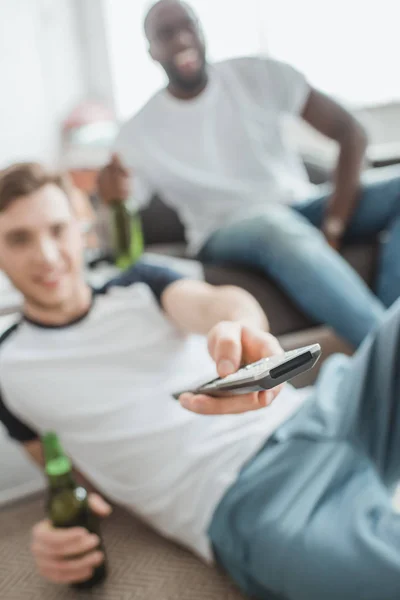 Closeup shot of remote in hand of man sitting with bottle of beer near friend — Stock Photo