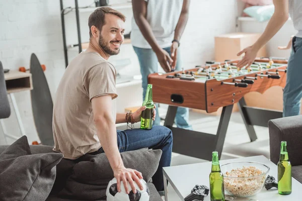 Sonriente joven con bola y cerveza sentado cerca de amigos jugando futbolín - foto de stock