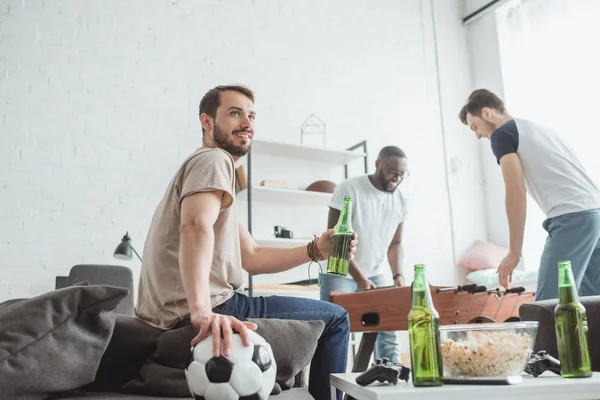 Baixo ângulo de visão do jovem com bola e cerveja sentado perto de amigos jogando futebol de mesa — Fotografia de Stock