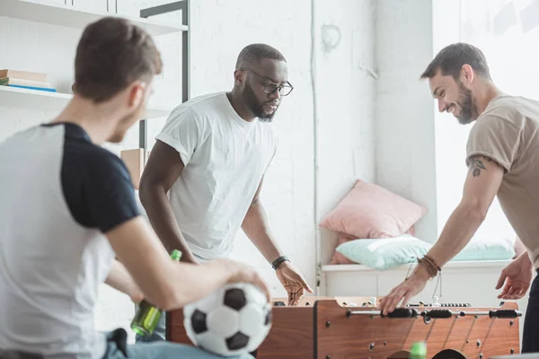 Vista posteriore dell'uomo con palla e birra guardando due amici multiculturali giocare a calcio balilla — Foto stock