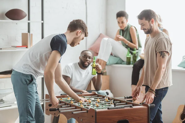 Side view of two young men playing table football while their friends watching with beer bottles — Stock Photo