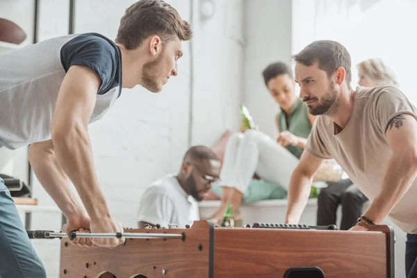 Young men with facial expression playing table football with friends sitting behind — Stock Photo