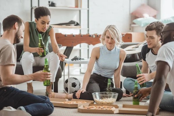 Mujer afroamericana señalando con el dedo a la pizza y amigos multiculturales de grupo sentados cerca en el suelo - foto de stock