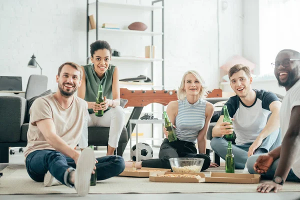 Smiling multiethnic friends sitting on floor with bottles of beer, pizza and popcorn — Stock Photo