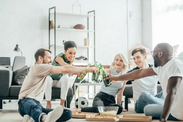 Happy multiethnic friends sitting on floor and clinking with bottles of beer — Stock Photo