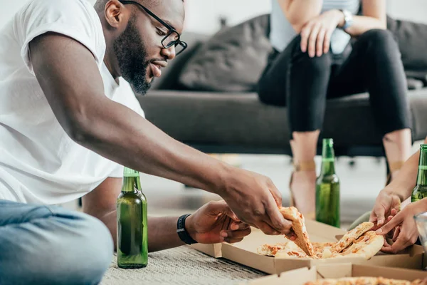 Homem americano africano elegante em óculos tomando pizza e sentado no chão com garrafas de cerveja — Fotografia de Stock