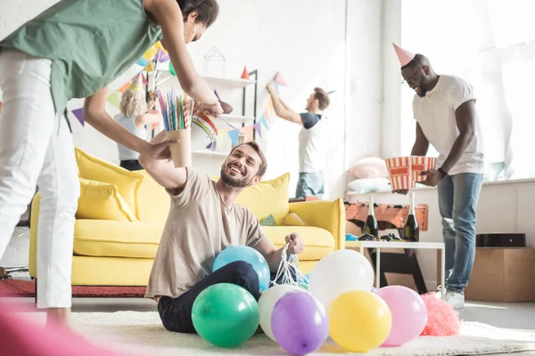 Vista frontale della donna afro-americana e sorridente giovane sul pavimento con palloncini ei loro amici decorazione stanza dietro — Foto stock