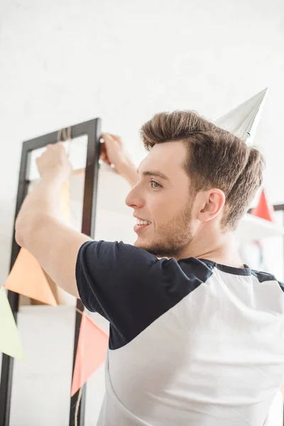 Smiling young man in party hat hanging up party garlands — Stock Photo