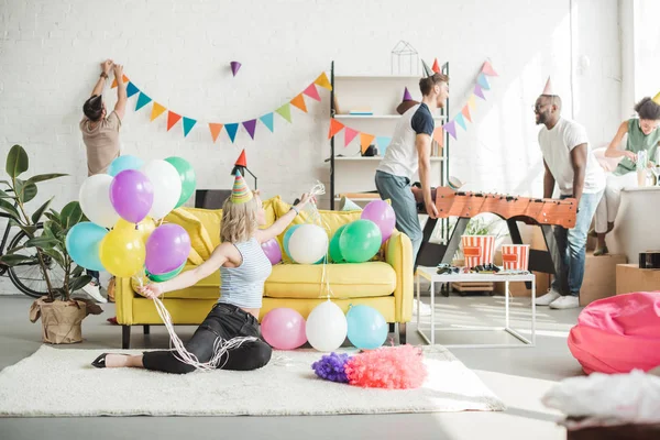 Young woman sitting on floor with colorful balloons and her friends decorating room behind — Stock Photo
