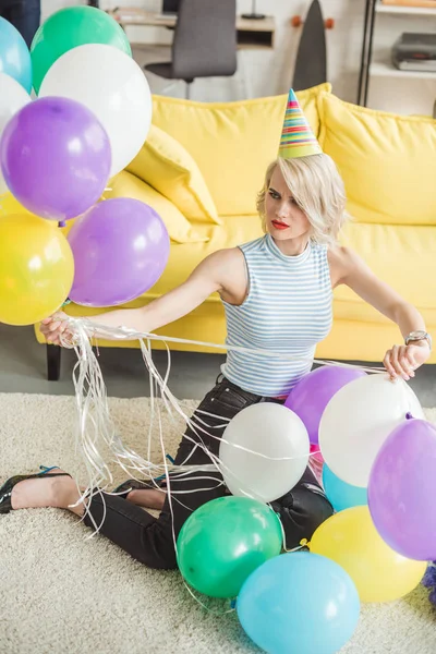 Young attractive woman in party hat siting on floor with colorful balloons — Stock Photo