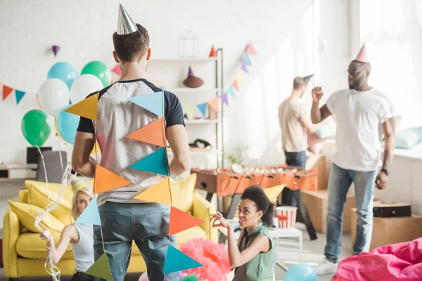 Vista trasera del joven envuelto en guirnaldas de fiesta y amigos de pie detrás de la habitación - foto de stock