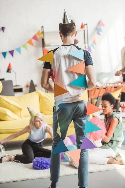 Vue arrière du jeune homme enveloppé dans des guirlandes de fête et de deux femmes multiculturelles assises sur le sol avec décor — Photo de stock