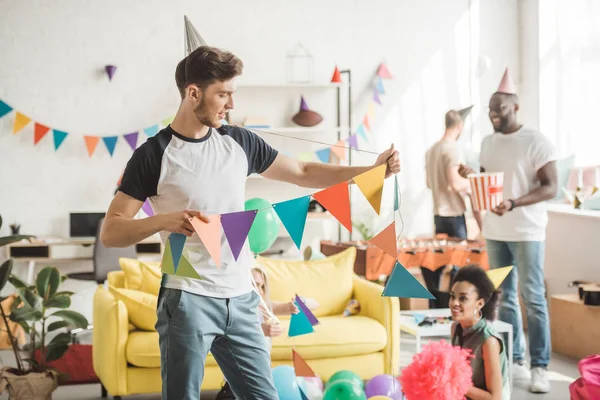 Jovem em chapéu de festa segurando corda com guirlandas de festa e amigos de pé atrás no quarto — Fotografia de Stock