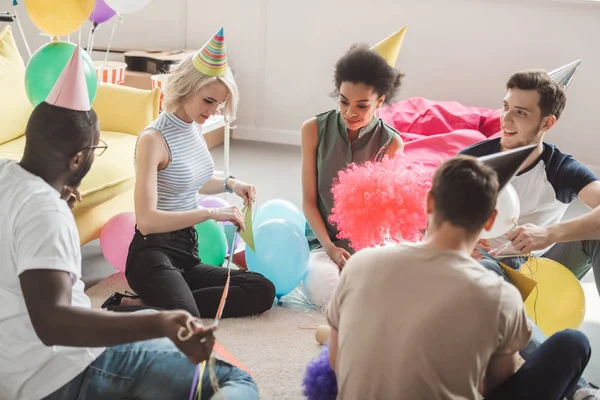 Grupo de jovens amigos multiculturais em chapéus de festa sentado no chão com balões em quarto decorado — Fotografia de Stock