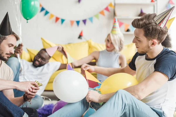 Amigos multiétnicos en sombreros de fiesta sentados en el suelo con globos en la habitación decorada - foto de stock