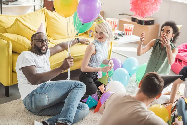 Amis multiculturels dans des chapeaux de fête assis sur le sol avec des ballons dans la chambre décorée — Photo de stock