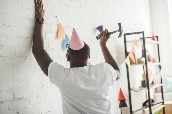 Young african american man with hammer nailing up string with party garland on brick wall — Stock Photo