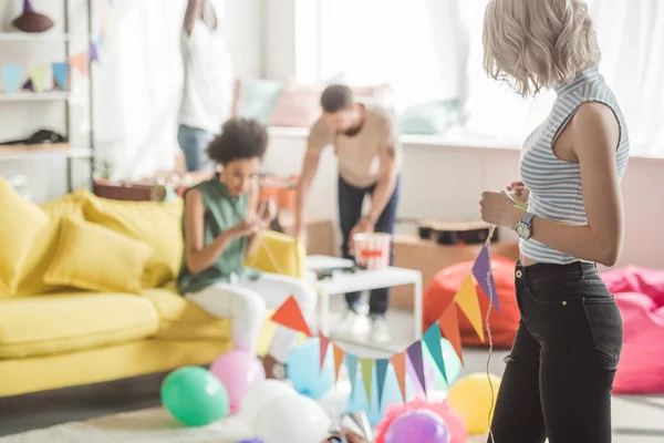 Zwei junge Frauen, die eine Kette mit bunten Partygirlanden in der Hand halten und Freunde, die dahinter stehen — Stockfoto